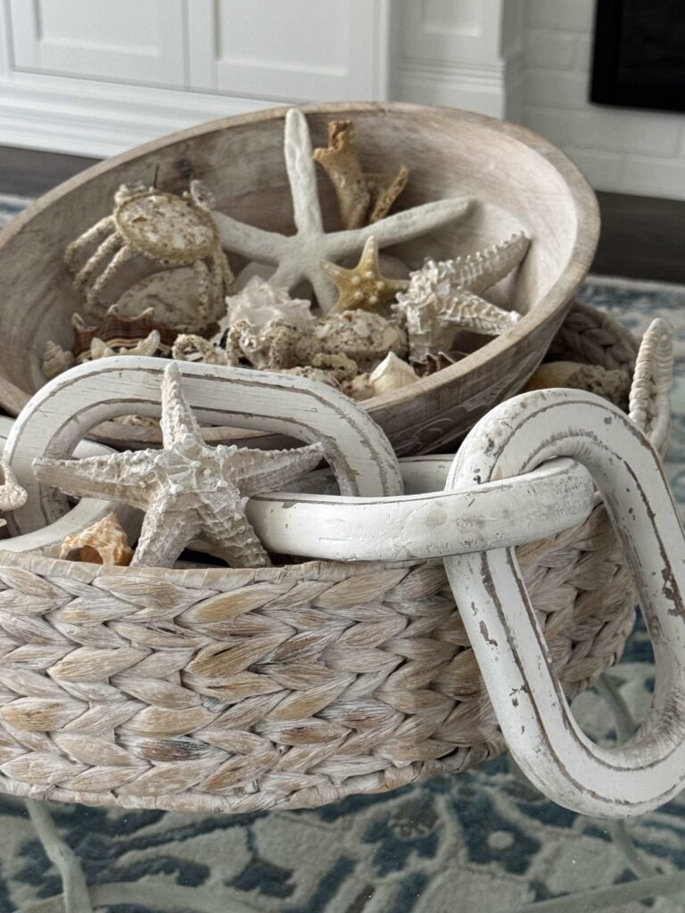 A woven basket holds a wooden bowl filled with starfish and seashells, offering effortless summer decor ideas. Large decorative wooden links rest on the basket's edge, completing the coastal-inspired scene that sits on a blue patterned surface.