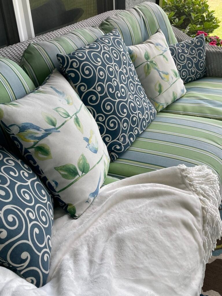 A green-striped outdoor sofa with blue and white swirl-patterned pillows, two pillows featuring a leaf and bird design, and a white blanket draped across the seat creates a cozy retreat. Green plants are visible in the background, inspiring ideas for how to hang flower pots to enhance your outdoor oasis.