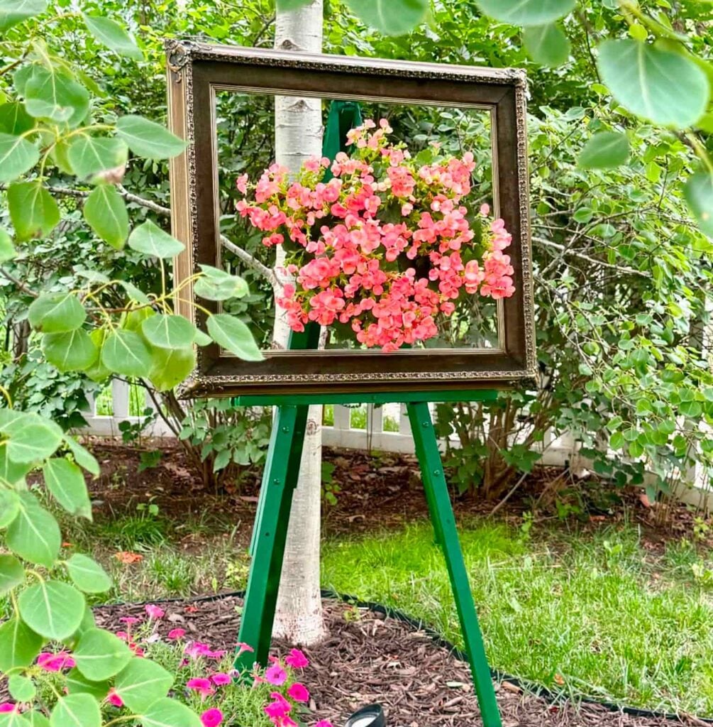 A vintage picture frame on a green easel stands outdoors, framing pink flowers and greenery, creating a living artwork effect in a garden setting—an inspiring display for anyone interested in creative gardening ideas such as how to hang flower pots.