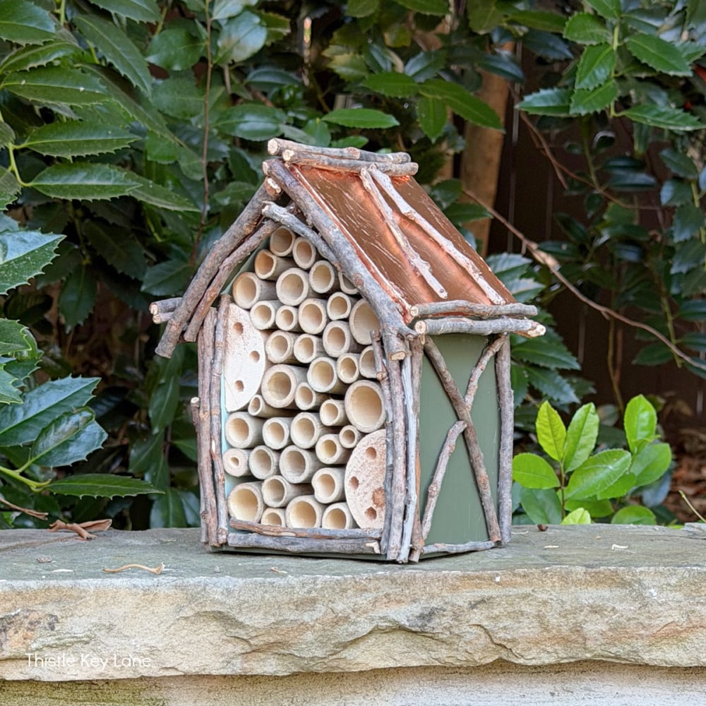 A small, rustic insect hotel made of twigs, bamboo tubes, and wood sits on a stone surface outdoors, surrounded by green leafy plants—an inviting spot for pollinators in any garden. Much like learning how to hang flower pots to enhance your outdoor space, adding features like this insect hotel can bring both beauty and function to your garden sanctuary.