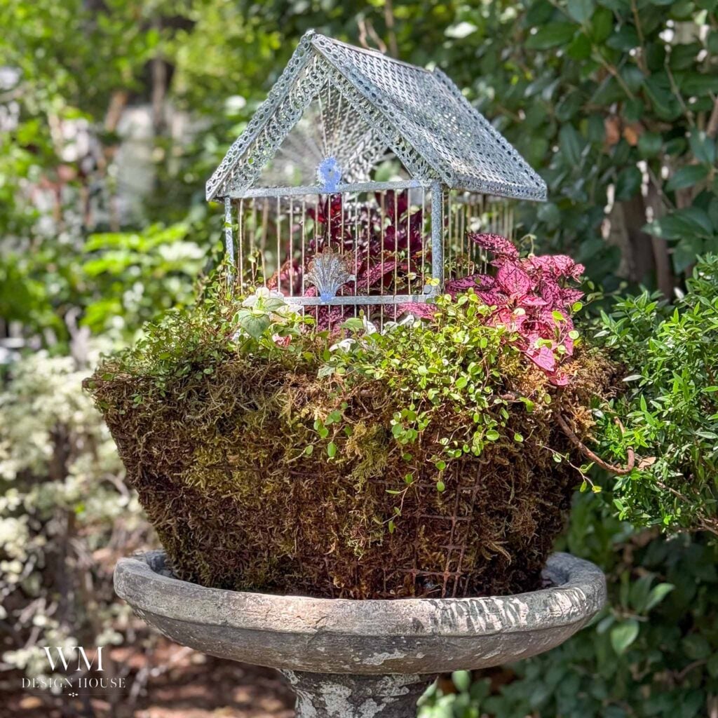 A decorative metal birdcage sits atop a moss-covered planter with pink and green plants, displayed on a stone pedestal outdoors, surrounded by lush greenery—a beautiful alternative if you’re seeking inspiration on how to hang flower pots in your garden.