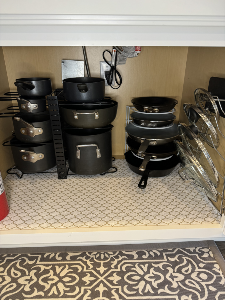 Neatly organized kitchen cabinet with stacked pots, pans, and lids. Pots and pans are black with metal handles, and lids are arranged vertically on the right. A fire extinguisher is visible on the left.