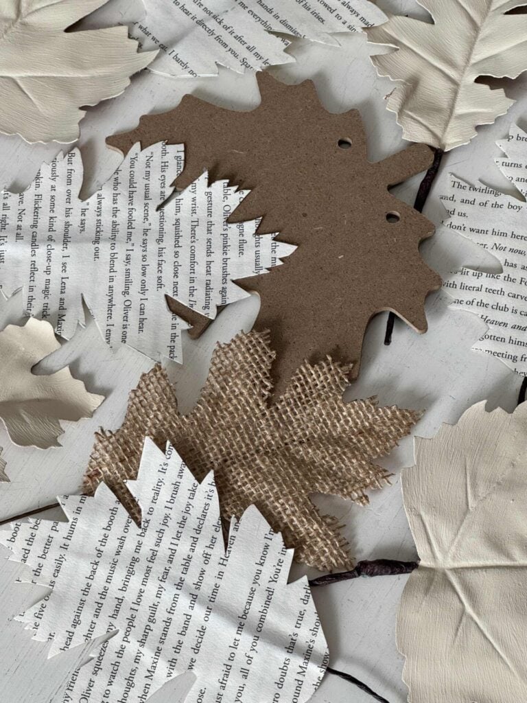 Assorted paper and fabric leaves, some made from book pages, burlap, cardboard, and cream-colored material, arranged flat on a surface.