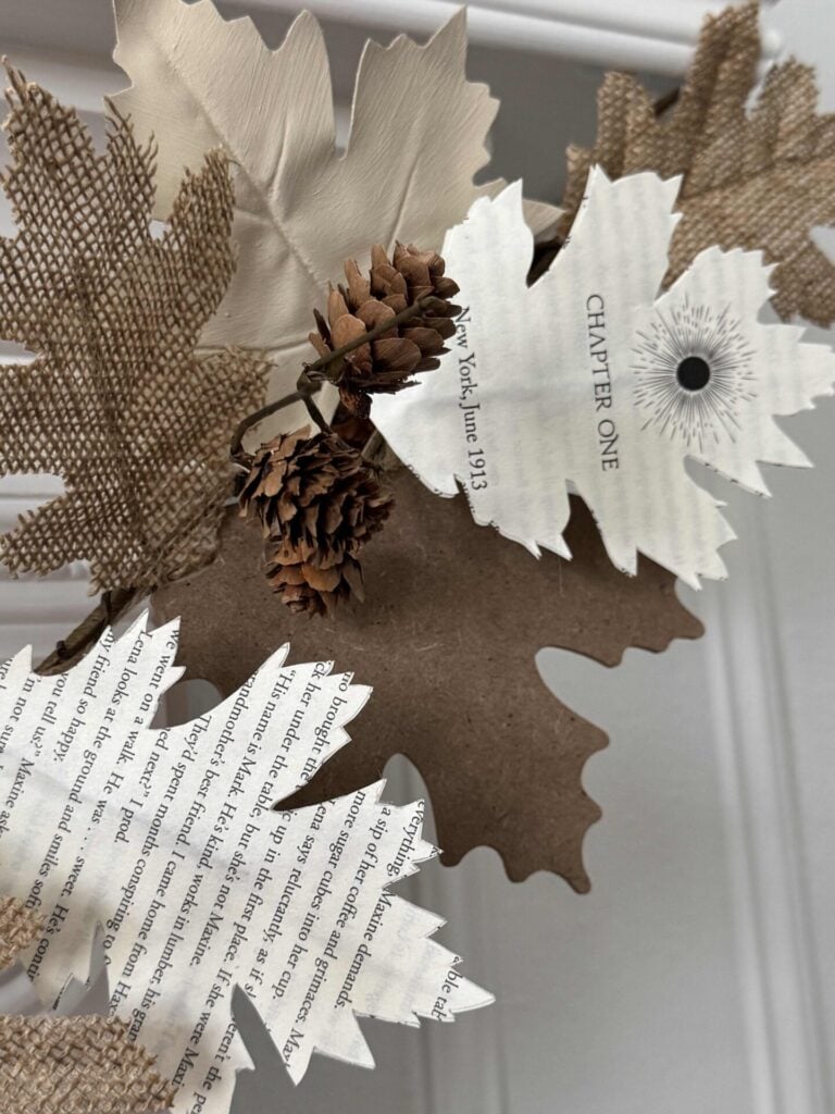Paper, burlap, and cardboard leaves, some made from book pages, are arranged with small pinecones to create a decorative autumn wreath—a charming example of DIY fall decor ideas. The visible book page says CHAPTER ONE.