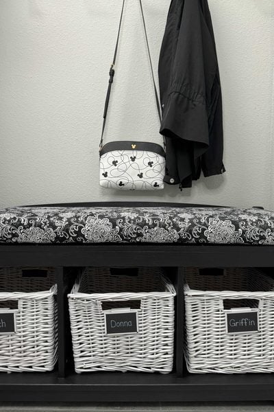 A black bench with a patterned cushion sits above three white baskets labeled Rich, Donna, and Griffin, showcasing stylish home organization products. Above the bench, a black coat and a white floral purse hang on a gray wall.
