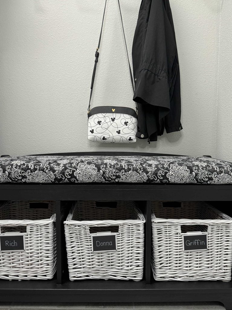 A black coat and white floral purse hang above a black bench with a patterned cushion, showcasing stylish home organization products. Below are three white baskets labeled Rich, Donna, and Griffin.