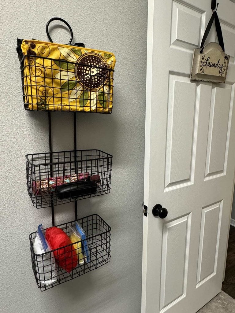 Three black wire baskets, part of a stylish collection of home organization products, are mounted on a wall by a white door. The top basket holds a yellow sunflower bag; the middle has folded items, and the bottom holds towels. A Laundry sign hangs on the door.