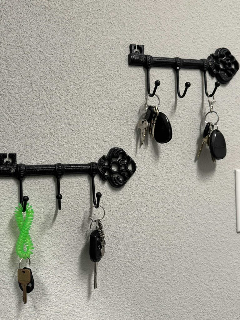 Two black, vintage-style key-shaped wall hooks—perfect examples of stylish home organization products—hold sets of keys, car fobs, and a green spiral wrist keychain, mounted on a light gray, textured wall. Part of a white light switch is visible on the right.