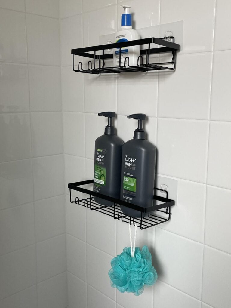Two black shower shelves on a white tiled wall hold lotion, two Dove Men+Care bottles, and a teal shower pouf hanging from the lower shelf.