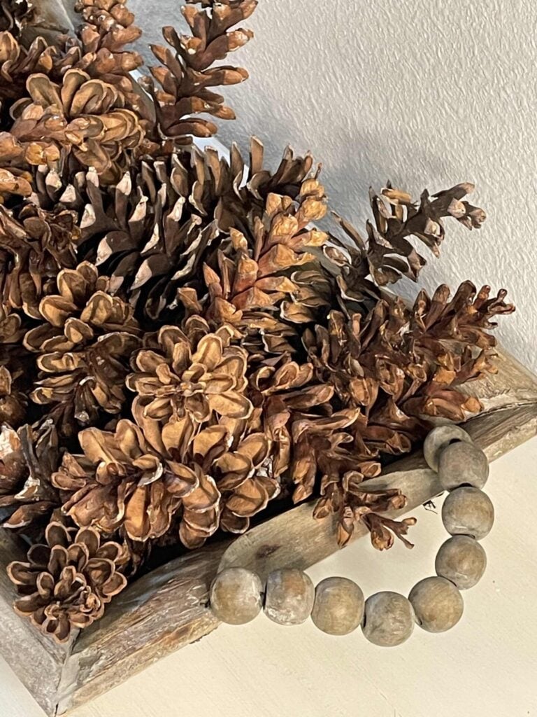 A wooden tray filled with brown pinecones sits on a light surface. The tray features a handle made of round wooden beads, adding a rustic touch perfect for seasonal displays.