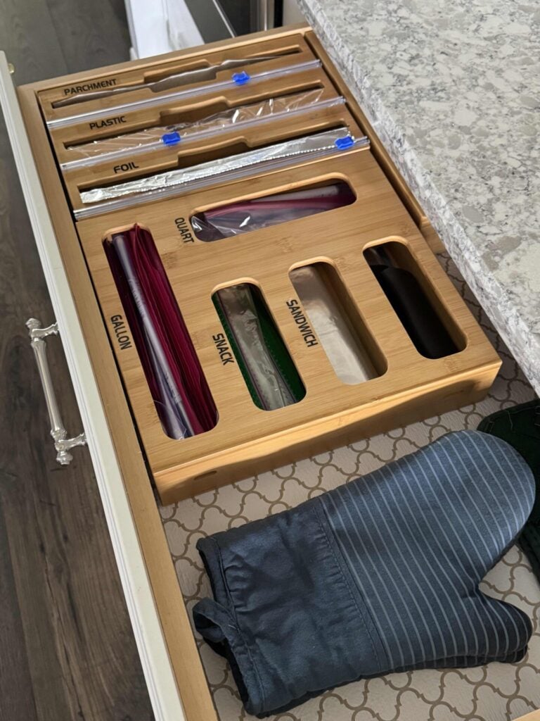 Open kitchen drawer with wooden organizer holding parchment, plastic wrap, foil, and various food storage bags (gallon, quart, snack, sandwich). An oven mitt rests beside the organizer on the drawer liner.