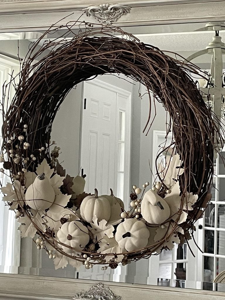 A rustic wreath made of twigs, decorated with white pumpkins, cream berries, and beige leaves, hangs on a mirror in a light-filled room.