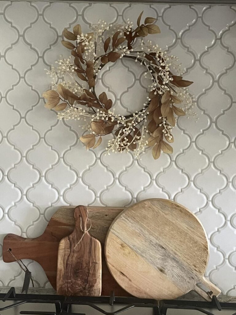 A decorative wreath with leaves and white berries, inspired by DIY fall decor ideas, hangs above three wooden cutting boards of different shapes, set against a white tiled backsplash.