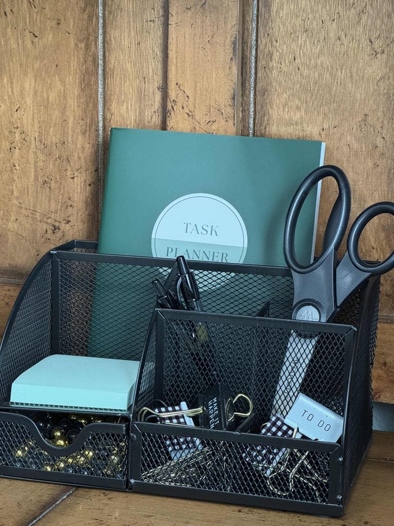 A black metal desk organizer, one of the most practical home organization products, holds scissors, pens, binder clips, a notepad, paper clips, and a TO DO label, with a green task planner propped behind it on a wooden surface.