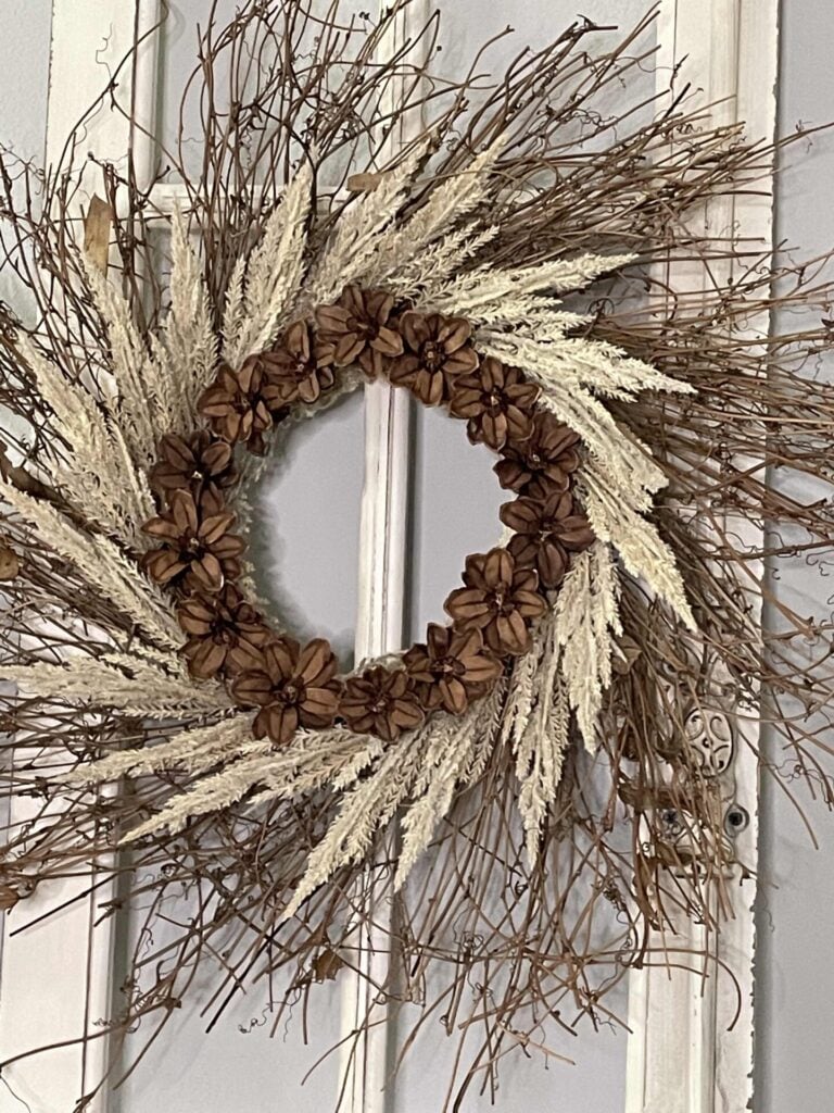 A rustic wreath made of dried twigs, beige grasses, and brown seed pods hangs on a white wooden door with vertical panels and an ornate metal keyhole plate, offering charming inspiration for DIY fall decor ideas.