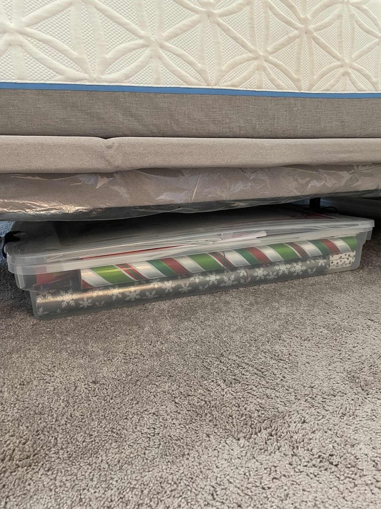 A clear plastic storage bin filled with rolled gift wrap is tucked under a bed with a gray fabric frame, resting on a beige carpet.