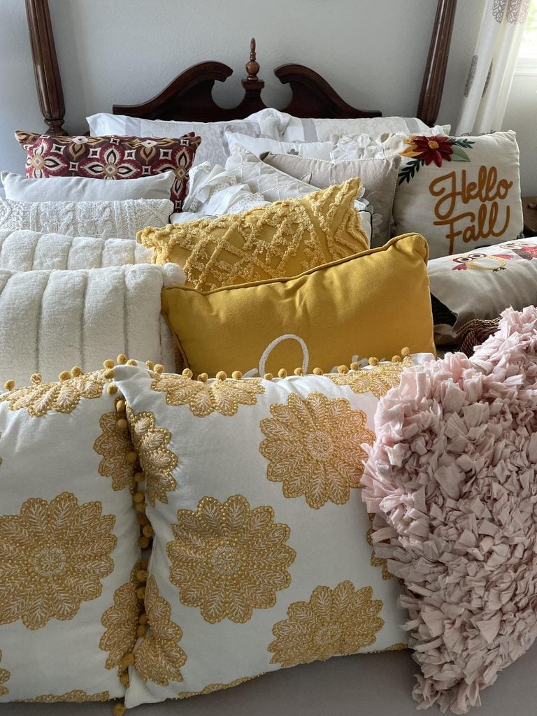 A bed is decorated with an assortment of pillows in yellow, white, pink, and patterned designs. One pillow says Hello Fall and another has a large letter O, creating a cozy and festive look that beautifully complements your favorite home organization products.