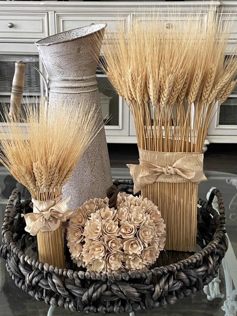 A woven tray holds a decorative pitcher, two bundles of dried wheat tied with ribbon, and a flower-covered pumpkin—an inviting arrangement that inspires DIY fall decor ideas—displayed on a glass table in front of white cabinets.