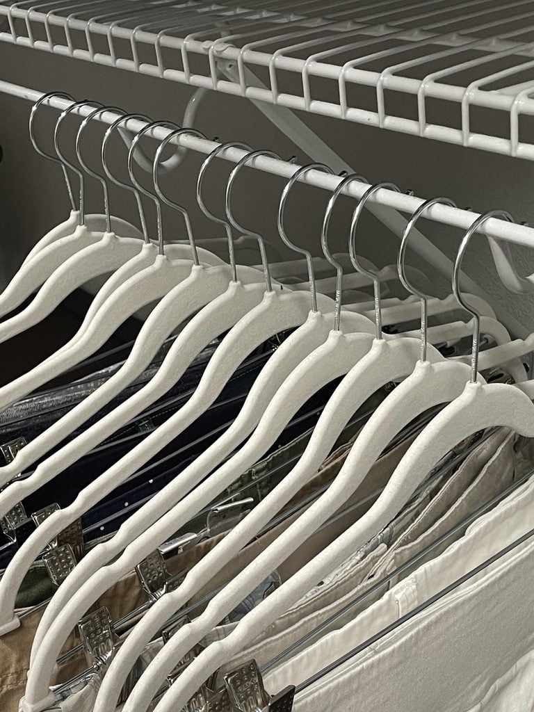 Several white velvet hangers are neatly arranged on a metal closet rod, holding a variety of clothes. A wire shelf is positioned above the hangers.