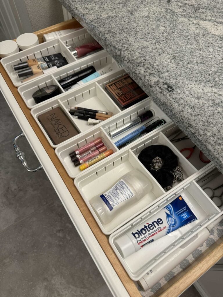 An organized bathroom drawer with white dividers holds makeup, brushes, toothpaste, lotion, hair ties, nail clippers, and other toiletries beneath a granite countertop.
