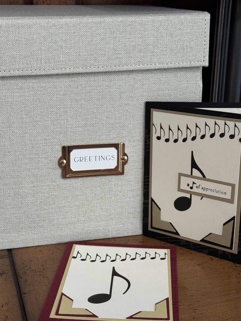 A gray box labeled GREETINGS—perfect for keeping your special cards tidy—sits on a wooden surface next to two music-themed greeting cards featuring musical notes and staff designs in black, beige, and maroon colors. This charming setup doubles as both decor and one of your essential home organization products.