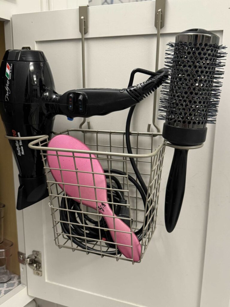 A wire basket hangs on a cabinet door and holds a black hair dryer, a round brush with metal bristles, and a pink hairbrush. The basket is mounted inside a white cabinet.
