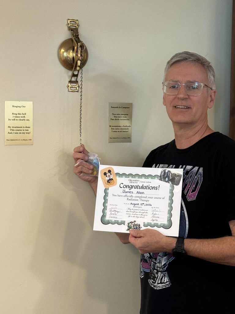 A man stands by a wall-mounted brass bell, holding the bells chain and a certificate that says Congratulations! with his name and cartoon animal graphics. He is smiling and wearing glasses and a dark graphic t-shirt.