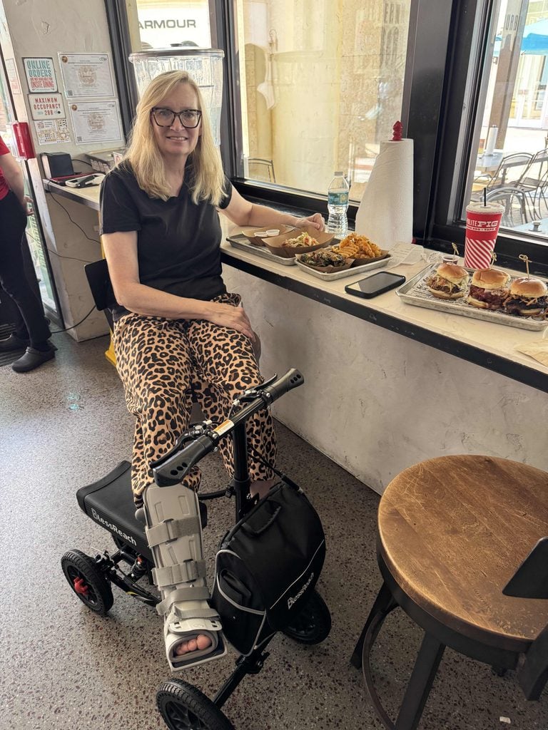 A woman with a leg brace sits on a knee scooter by a window counter, eating food. She wears glasses, a black shirt, and leopard-print pants. Food and drinks are on the counter beside her.