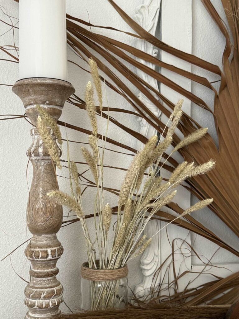 A rustic candle holder with a white candle stands next to a jar of dried grasses; dried palm leaves decorate the white textured background.