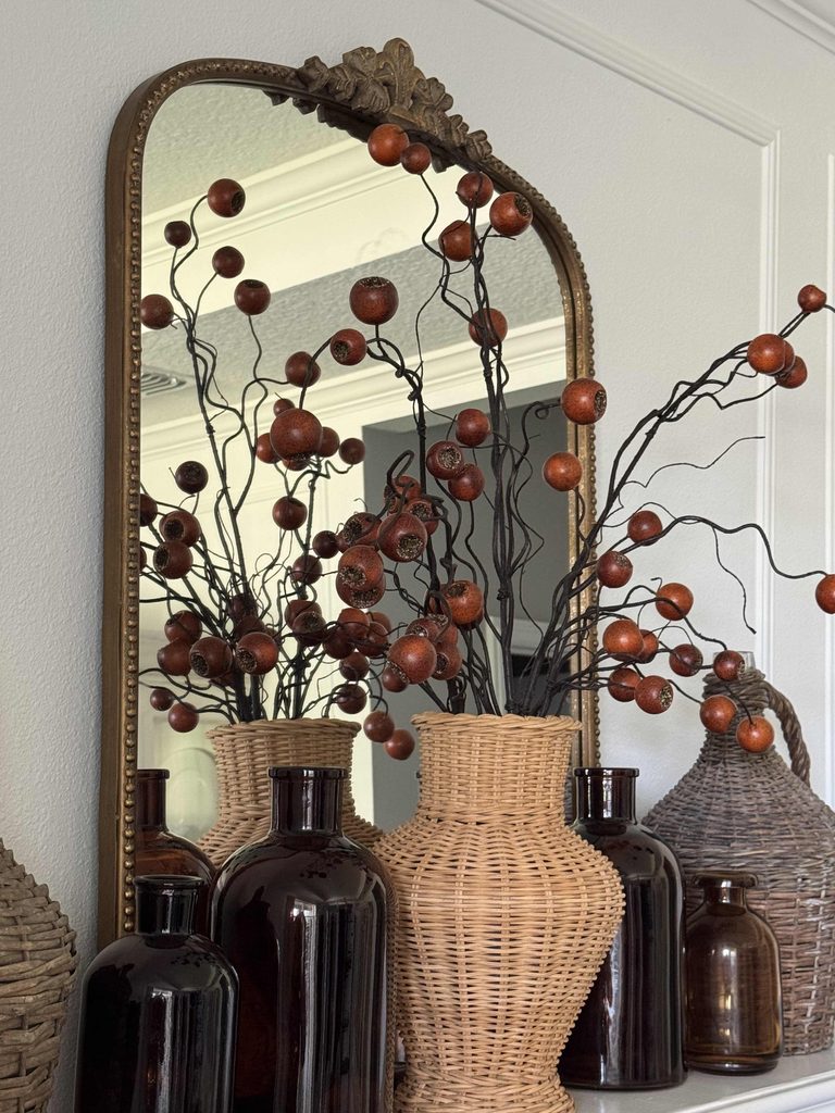 A decorative mirror reflects branches with orange berries arranged in brown glass and woven wicker vases, set on a white mantel against a white wall with crown molding.
