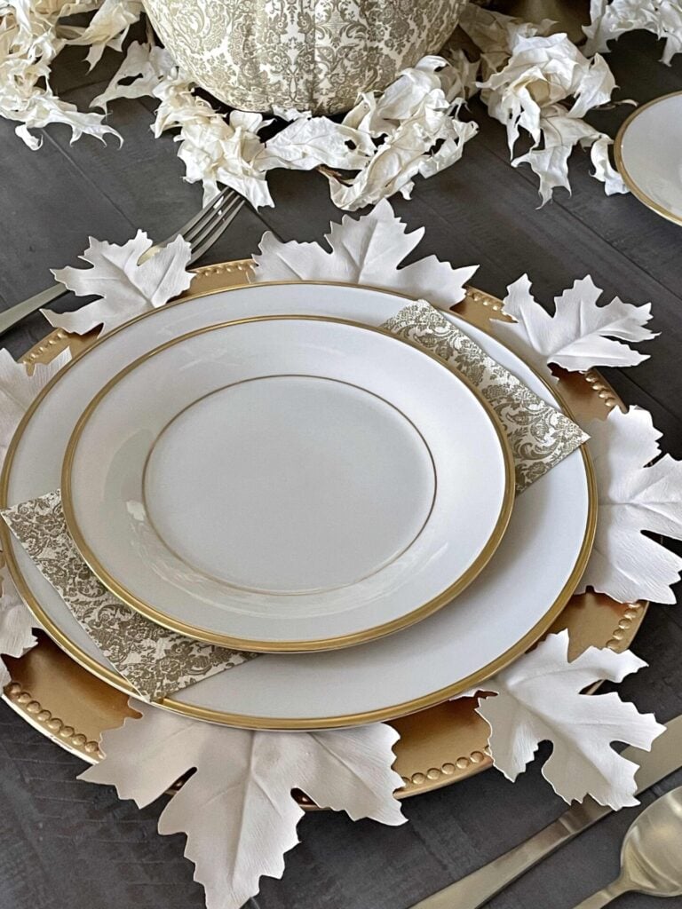 A fall-themed table setting with white plates on a gold charger, decorated with white faux leaves and patterned napkins, arranged neatly on a wooden table—perfect inspiration for your DIY fall decor ideas.