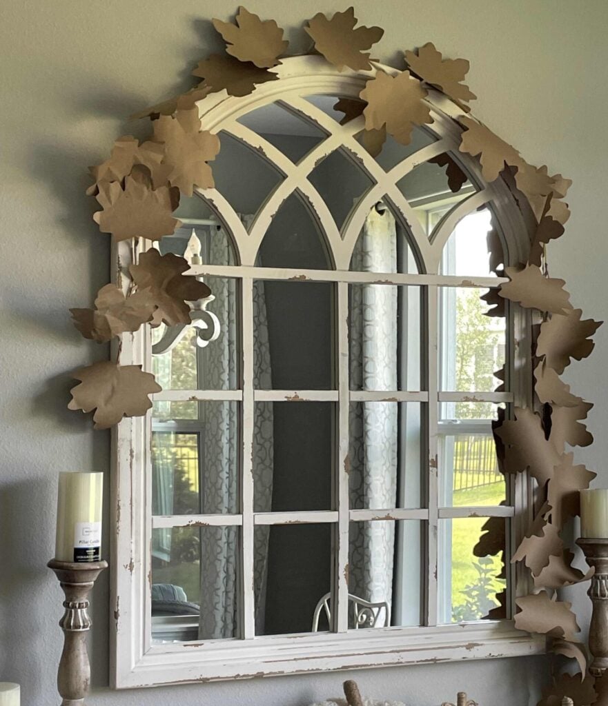 A white arched mirror with windowpane design is decorated with brown paper leaves along the top and side. Candles and light from a nearby window are beautifully reflected in the mirror.