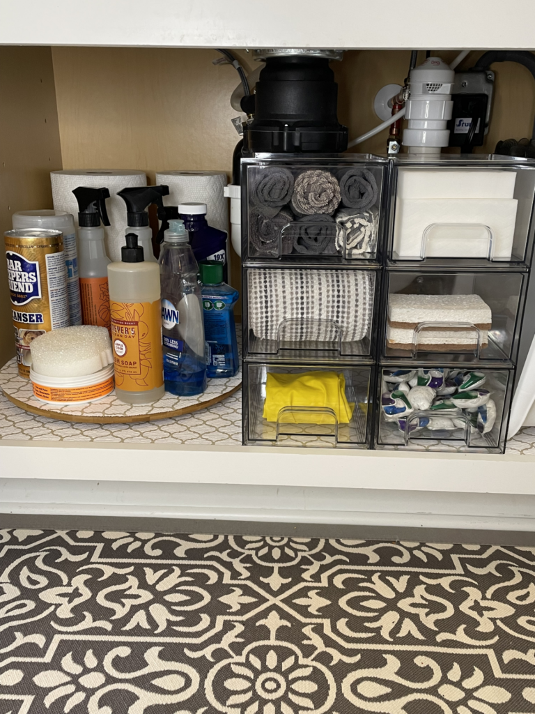 Organized cabinet under a sink with cleaning supplies, towels, and detergents neatly stored in clear drawers and containers—showcasing the effectiveness of home organization products. A patterned rug is on the floor in front.