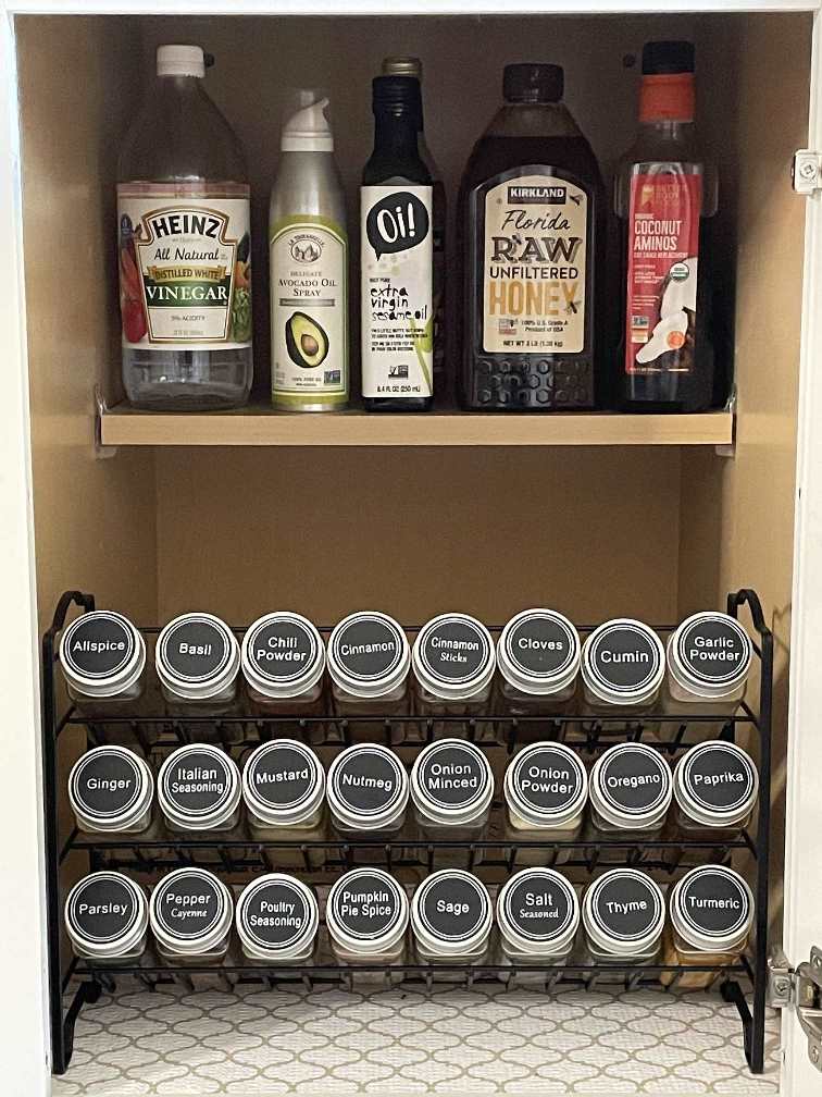 A neatly organized spice rack, featuring top home organization products, sits below a shelf holding vinegar, oil, honey, aminos, and coconut oil. Labeled spice jars are arranged in tidy rows, all inside a white kitchen cabinet.