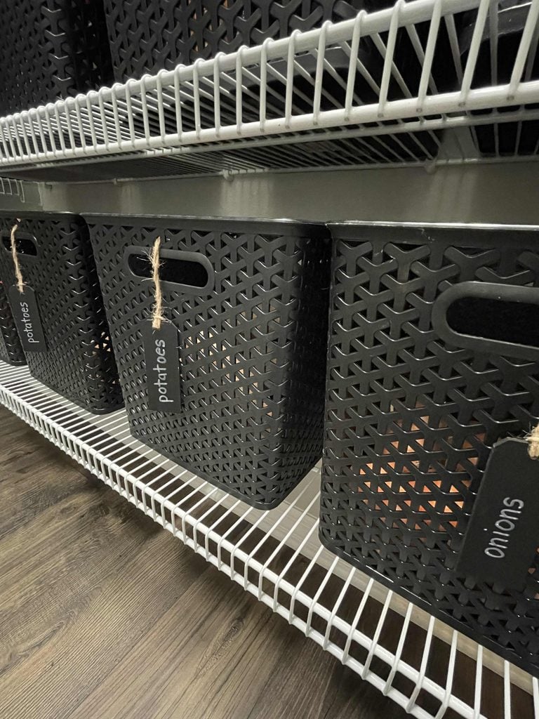 Black plastic baskets with labels for potatoes and onions sit on white wire shelves, offering a tidy storage solution among your favorite home organization products. The floor is wooden, and the baskets feature convenient handles and tags attached with string.