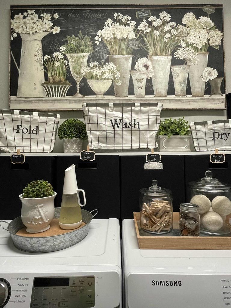 Laundry room featuring home organization products like labeled baskets for Fold, Wash, and Dry, jars holding clothespins and detergent, decorative plants, and a floral-themed wall art above the shelves. Washing machine and dryer are below.