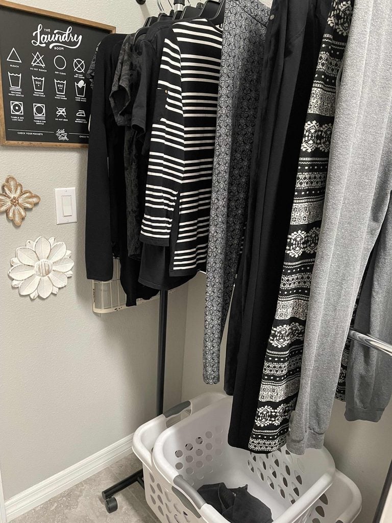 Clothes hang on a rack next to a white laundry basket and a wall decorated with laundry symbols and flowers; most clothes are black with striped or patterned designs, showcasing practical home organization products for a tidy, stylish space.