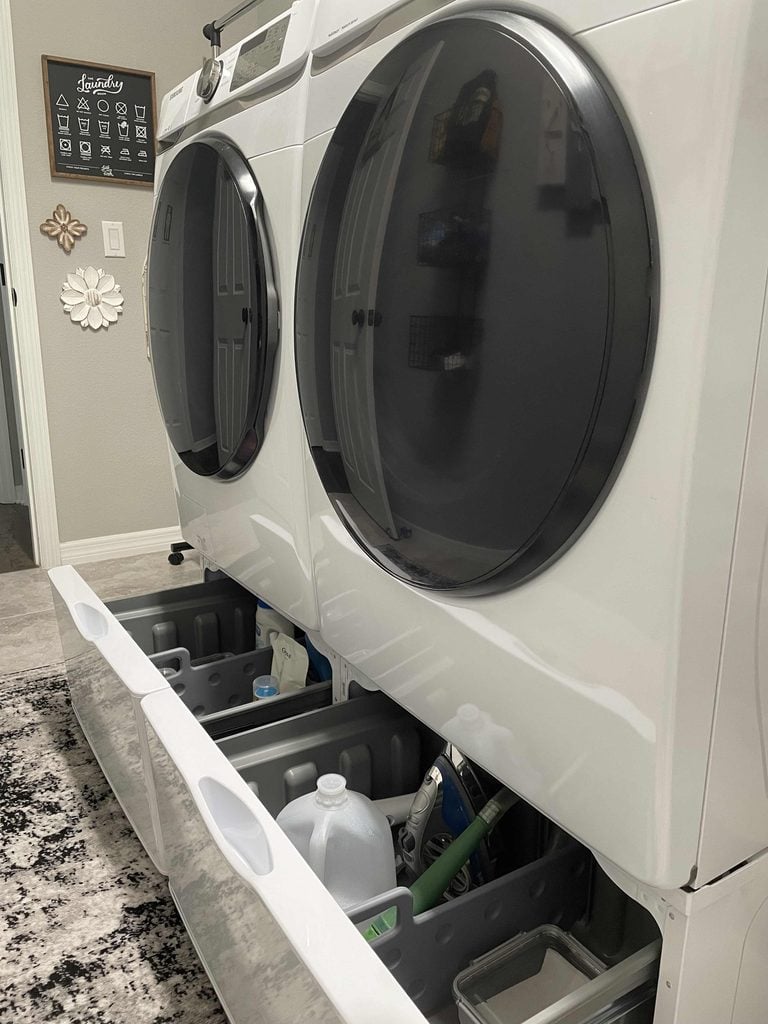 Front-load washer and dryer with bottom drawers open, revealing cleaning supplies and containers stored inside—perfect for showcasing your favorite home organization products. Laundry room decor and a wall sign are visible in the background.