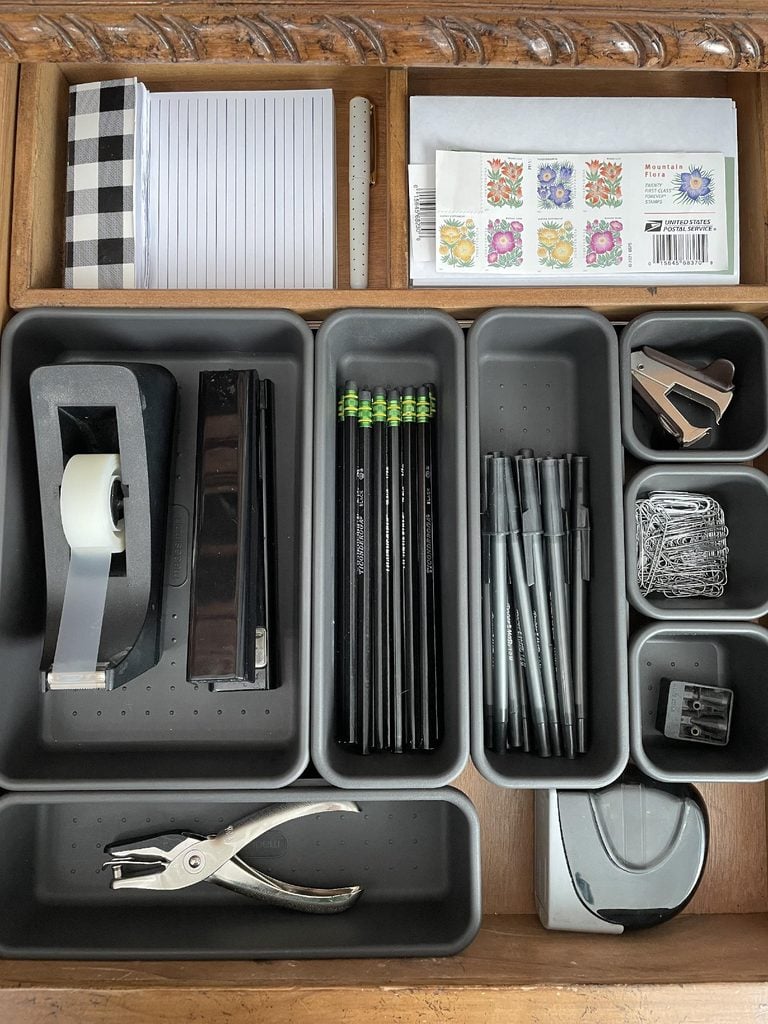 Drawer with organized office supplies: notepad, stamps, tape, stapler, pencils, pens, paper clips, binder clips, pencil sharpener, hole punch, and tape dispenser arranged in gray trays—an ideal setup for anyone seeking effective home organization products.