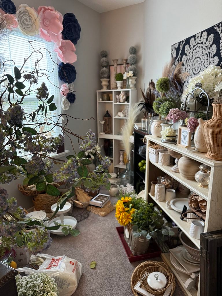 A cozy room filled with shelves, artificial flowers, home organization products, wicker baskets, vases, and charming décor. Large paper flowers hang by the window as sunlight filters in, casting a soft glow over the eclectic blend of decorative items.