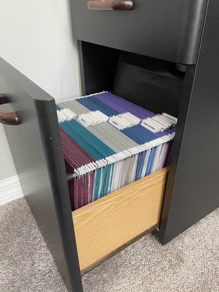 An open black filing cabinet drawer filled with organized hanging file folders in various colors, each labeled with white tabs. Showcasing the effectiveness of home organization products, the cabinet sits on a carpeted floor against a white wall.