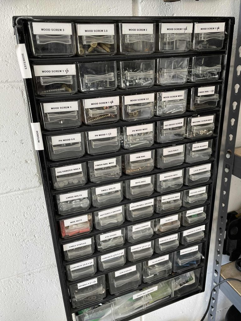 A black plastic organizer with many small labeled drawers, each containing various screws, nails, and hardware, is mounted on a white wall in a workshop or garage—demonstrating the effectiveness of quality home organization products.