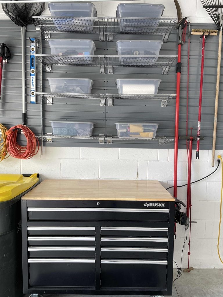 A garage wall with a Husky tool chest, four wire shelves holding clear storage bins, and various tools including rakes and cords hanging on either side showcases the effectiveness of quality home organization products.