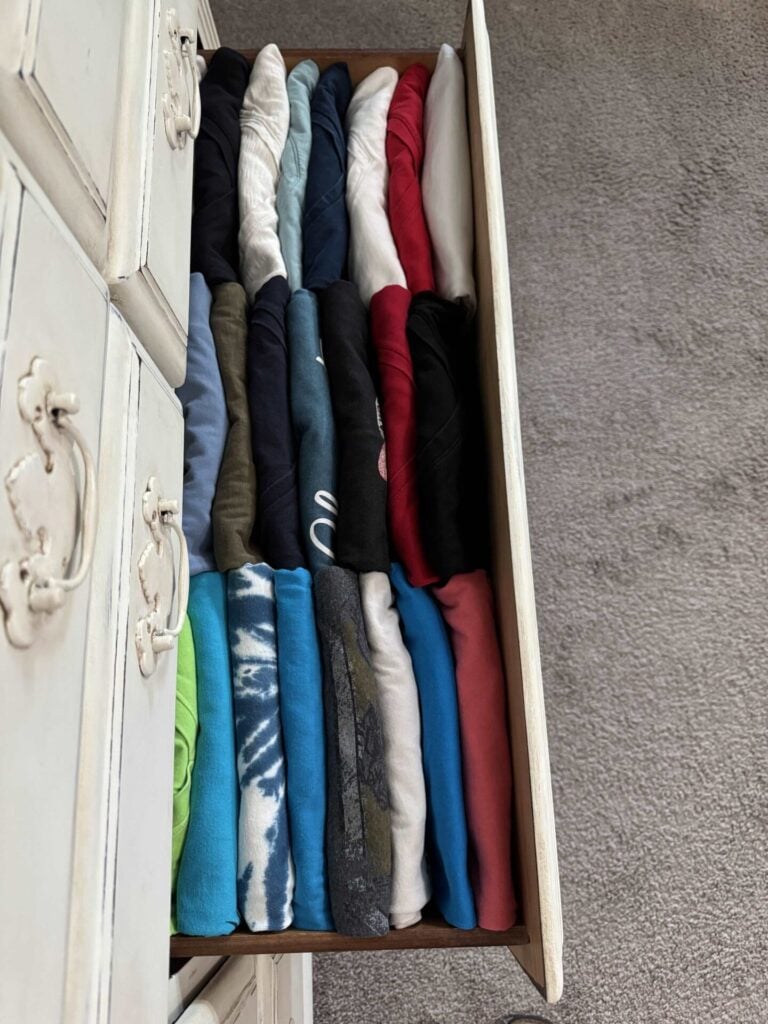 An open dresser drawer neatly filled with folded t-shirts arranged vertically in rows, showing various colors and patterns. The dresser is white and vintage-style, next to a light gray carpeted floor.