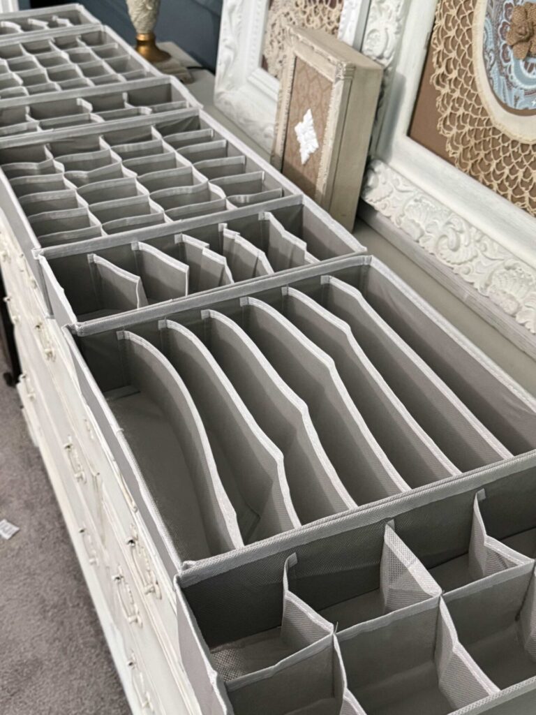 Gray fabric drawer organizers with multiple compartments are arranged on top of a white dresser. Decorative wall art and frames can be seen in the background.