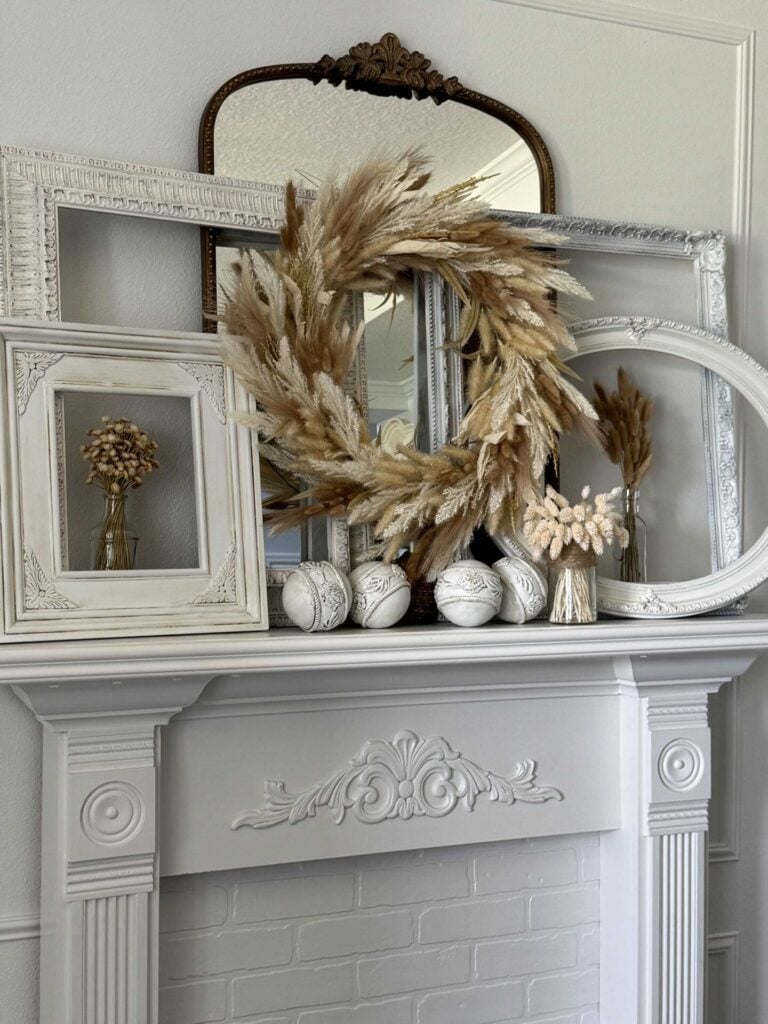 A white mantel decorated with empty picture frames, a pampas grass wreath, white decorative balls, and small vases with dried flowers, complemented by a vintage mirror in the background.
