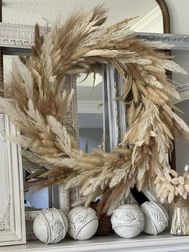 A large pampas grass wreath is displayed on a mantel with decorative white balls and ornate, layered mirrors in the background. Perfect for those seeking DIY fall decor ideas, the arrangement has a neutral, natural color palette and a rustic, elegant style.