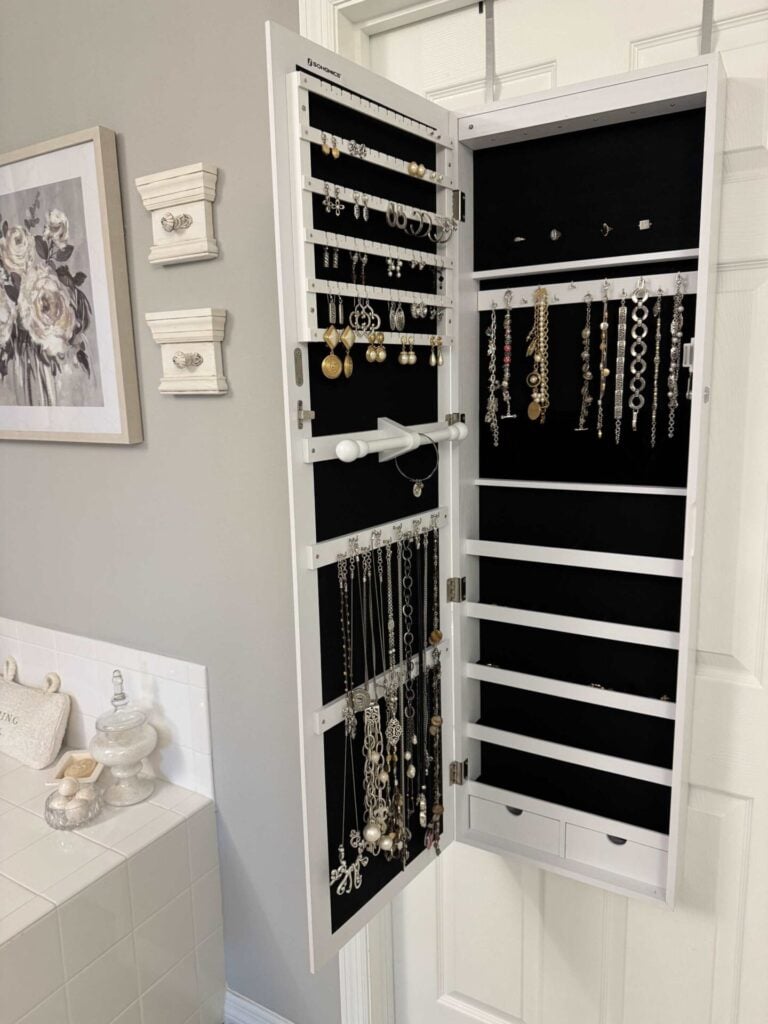 A white wall-mounted jewelry organizer, one of the must-have home organization products, displays necklaces, bracelets, and earrings neatly arranged inside as it hangs on the back of a door in a gray bathroom with white accents and floral decor.