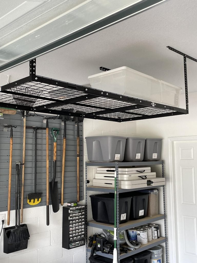 Overhead metal storage rack with a clear bin on top, shelving unit with labeled gray bins, and various tools like rakes and shovels hanging on a wall—this tidy garage showcases the power of home organization products.