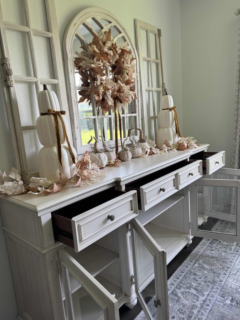 White sideboard with open drawers and doors, decorated with white pumpkins, autumn leaves, and a large wreath on a mirror. The decor has a fall theme with neutral colors in a bright, softly lit room.